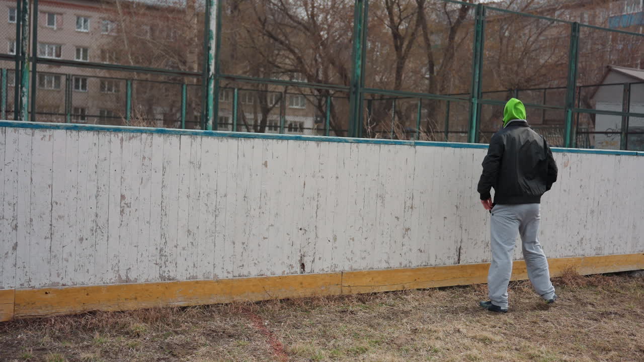 Hombre blanco trepa por encima de la barrera de la pista para recuperar un balón de fútbol, busca un punto de apoyo seguro en la cornisa de madera, pista urbana gris, esfuerzo solitario, resolución de problemas durante un partido informal, movimiento decidido