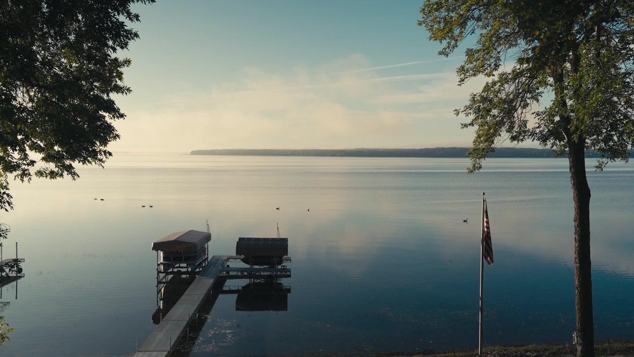 Calm lake at sunrise with geese swimming past a dock, boat lifts, trees, and an American flag reflecting on still water