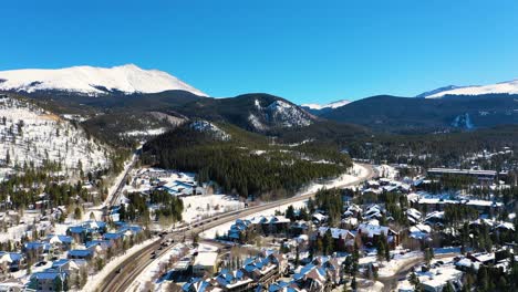 Vista-Aérea-Por-Drones-De-Casas-Y-Montañas-Cubiertas-De-Nieve-En-La-Hermosa-Ciudad-Montañosa-De-Breckenridge,-Colorado,-Con-Gente-Conduciendo-Por-La-Carretera-A-Través-De-Un-Bosque-De-Pinos