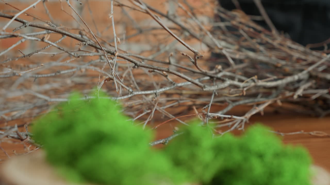 Female florist arranging dry twigs on wooden table with green moss in foreground, preparing natural craft decoration with eco friendly materials for handmade wreath in rustic workshop
