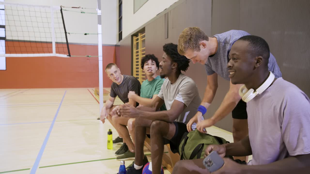 Diverses männliches Volleyballteam betritt die Turnhalle zum Training, sitzt auf der Bank, legt den Ball ab, checkt das Handy.