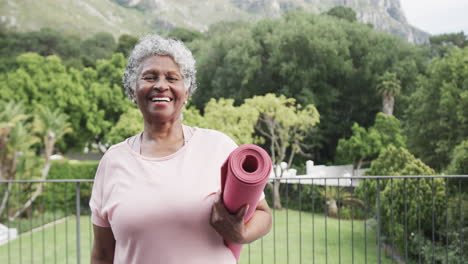 retrato de una feliz mujer afroamericana senior sosteniendo una alfombra de yoga en el balcón, espacio de copia, cámara lenta