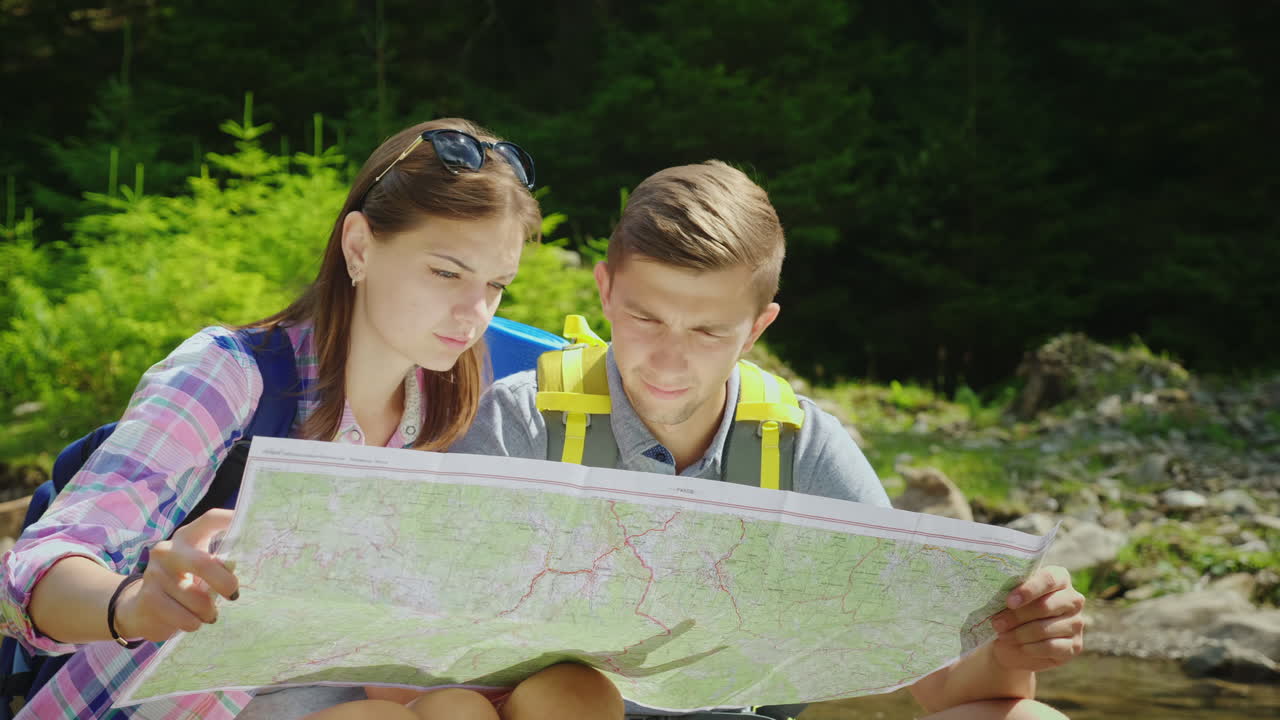 un par de jóvenes turistas están estudiando juntos un mapa que se sientan en un lugar pintoresco cerca de una montaña