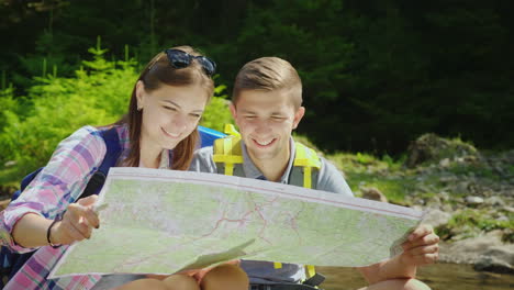 a young couple of tourists are studying together a map they sit in a picturesque place near a mounta