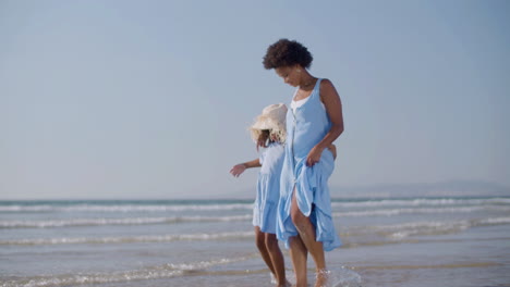 happy mother and daughter walking along seashore barefoot, splashing water under their feet and having fun together