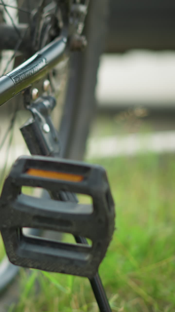 primer plano del pedal y la cadena de la bicicleta con la pierna de una persona preparándose para quitar el soporte, en un fondo verde exuberante, mostrando detalles intrincados de la bicicleta y un momento activo al aire libre en el entorno del parque