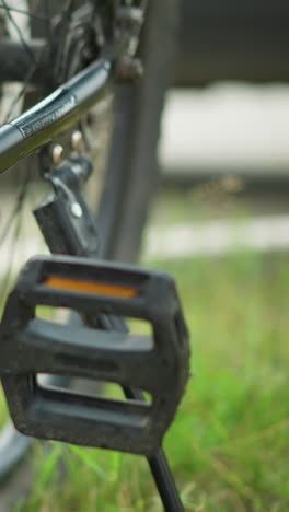 close-up do pedal e da cadeia da bicicleta com a perna de uma pessoa preparando-se para remover o suporte, definido contra um fundo verde exuberante, mostrando detalhes intrincados da bicicleta e momento ativo ao ar livre no cenário do parque