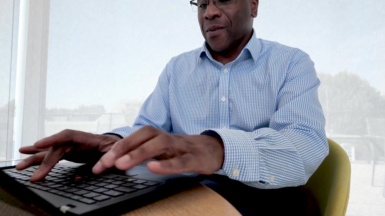 Black office employee in glasses types on a laptop at a tidy desk, with daylight from large windows and office supplies arranged nearby.
