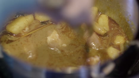 cooking malaysian chicken curry in a pot on the stove whilst stirring with a pair of metal tongs