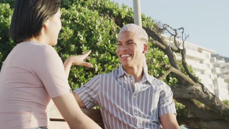 Happy-biracial-couple-sitting-on-bench-and-talking-on-promenade,-in-slow-motion