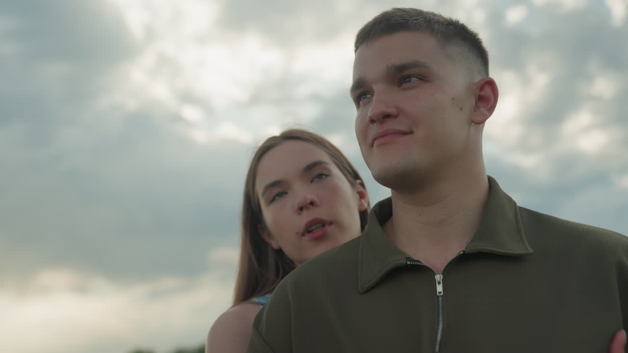 close up of woman holding man shoulder intimately as couple gaze into sky over rural field setting evoking romantic bond and serene connection under cloudy sky