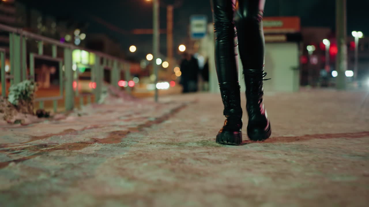 검은색 가죽 바지와 검은색 부츠를 입은 여성이 눈 인 거리에서 밤에 걸어가고, 흐릿한 도시 불빛으로