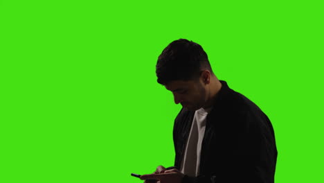 Young-Man-Writing-And-Sending-Text-Message-On-Mobile-Phone-Standing-Against-Green-Screen-Background-With-Low-Key-Lighting-1