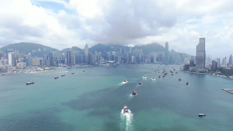 Convoy-of-local-Fishing-boats-causing-in-Hong-Kong-Victoria-bay,-with-city-skyline-in-the-horizon,-Aerial-view