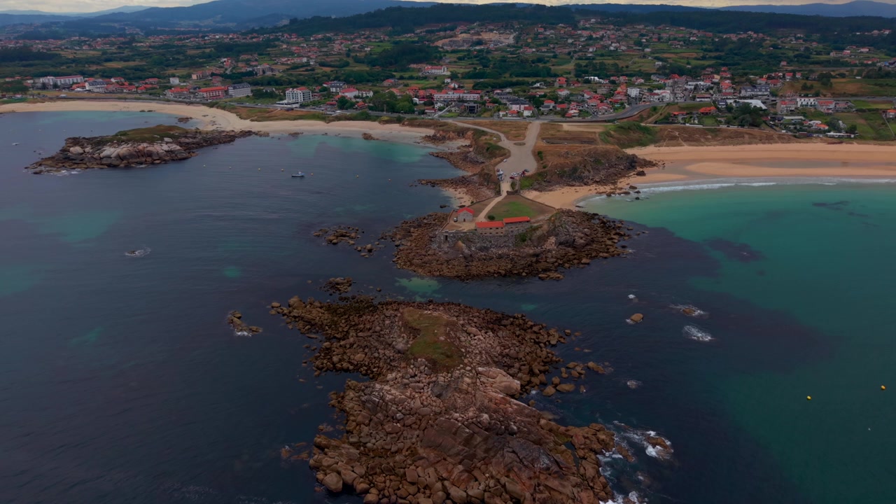 Droneudsigt fra luften af Nossa Senhora da Lanzada klippefyldte forbjerg med dets kapel, sandstrande og turkise vand på Galiciens kyst i Spanien.