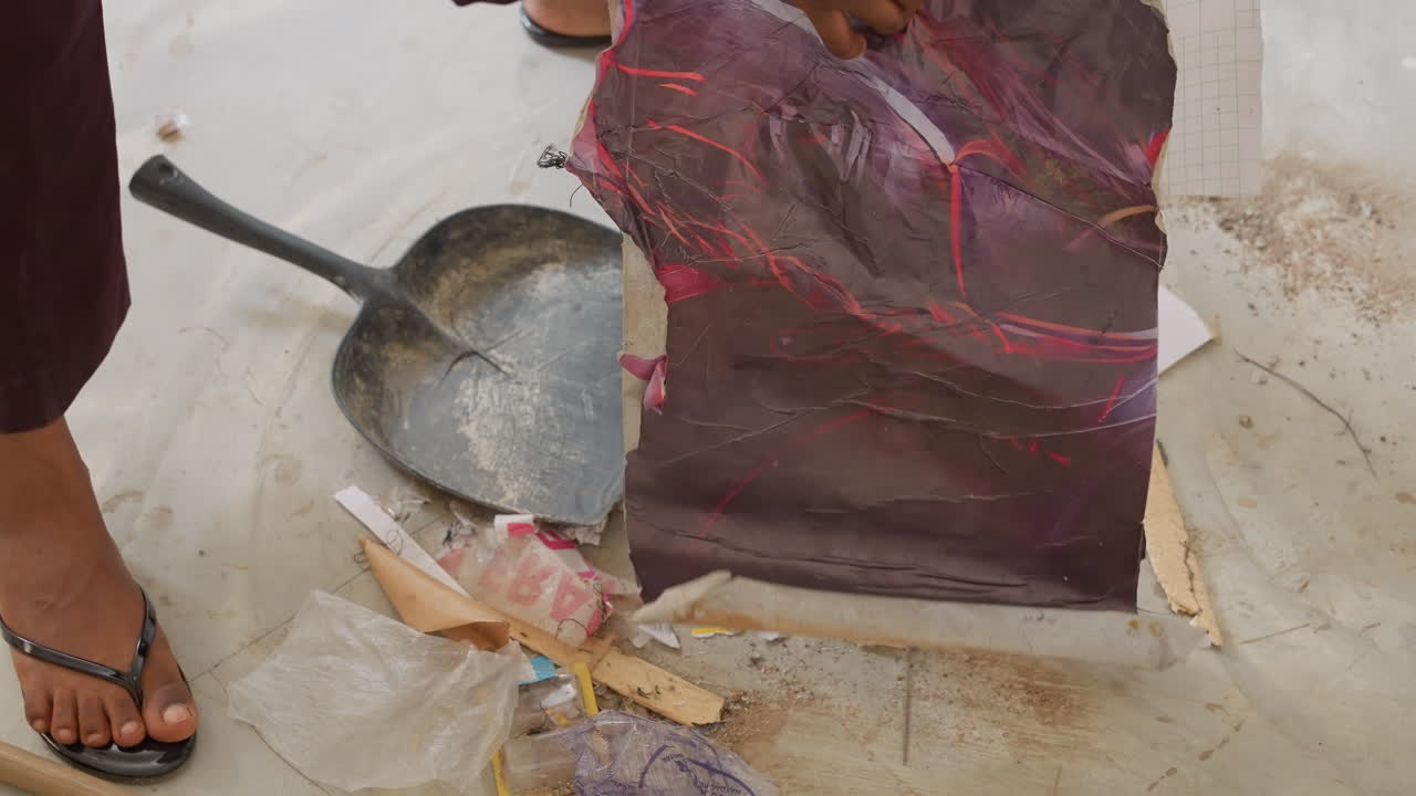 Proprietario di casa di colore che spazza detriti di casa e scarti di carta da parati, primo piano di sandali e mani che usano paletta e spazzola, sollevamento, piegatura e smaltimento attento di fogli strappati, scena di una pratica faccenda di ristrutturazione.