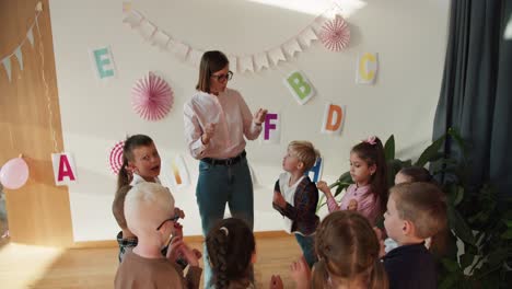 Niños-Felices-En-Edad-Preescolar-En-Un-Grupo-Haciendo-Un-Calentamiento-De-Muñecas-Antes-De-Escribir-Con-Su-Maestra,-Una-Niña-Con-Un-Peinado-Bob-Y-Anteojos-Con-Una-Camisa-Blanca-En-Un-Club-Especial-Para-Preparar-A-Los-Niños-Para-La-Escuela