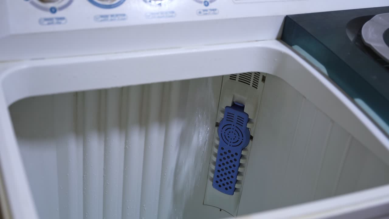 Close-up detail of clear water cascading down the interior wall of a washing machine. Focus on the blue plastic lint filter (trap) inside the white drum during the filling cycle