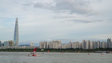 Gente-Haciendo-Deportes-Acuáticos-En-El-Río-Han-Al-Atardecer,-Edificio-Emblemático-De-La-Torre-Mundial-Lotte-En-El-Fondo-Del-Horizonte-De-Seúl,-Corea-Del-Sur