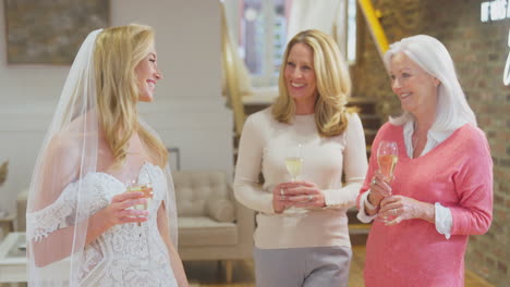 Three-Generation-Female-Family-Drink-Champagne-Adult-Daughter-Tries-On-Wedding-Dress-In-Bridal-Store