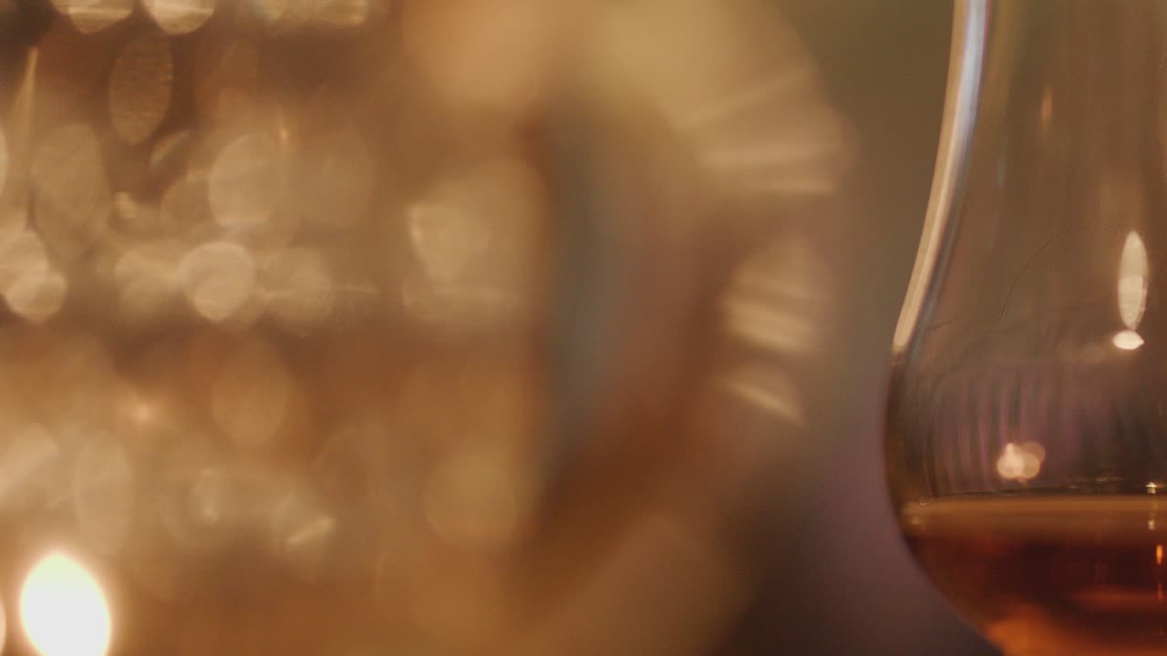 A close-up of whisky glasses on a table, softly lit by candlelight. The camera slowly shifts focus, creating a warm, blurred bokeh effect
