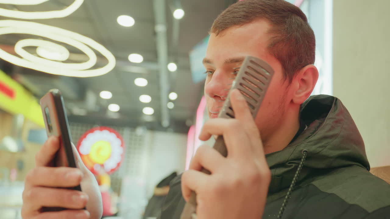 Uomo bianco che testa un microfono in un centro commerciale, con in mano il telefono, luci ad anello al neon sopra la testa, che pratica il canto, atmosfera da aspirante cantante, felpa con cappuccio casual, gesti di prova, ritratto ravvicinato, sfocatura della folla circostante.