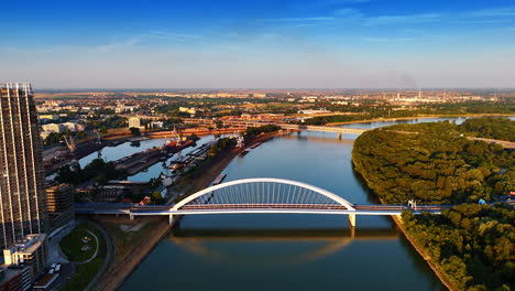 Arch of the beautiful Appolo Bridge reflects in the calm waterscape of the Danube River. Sunny view of the vast cityscape of Bratislava, Slovakia from drone