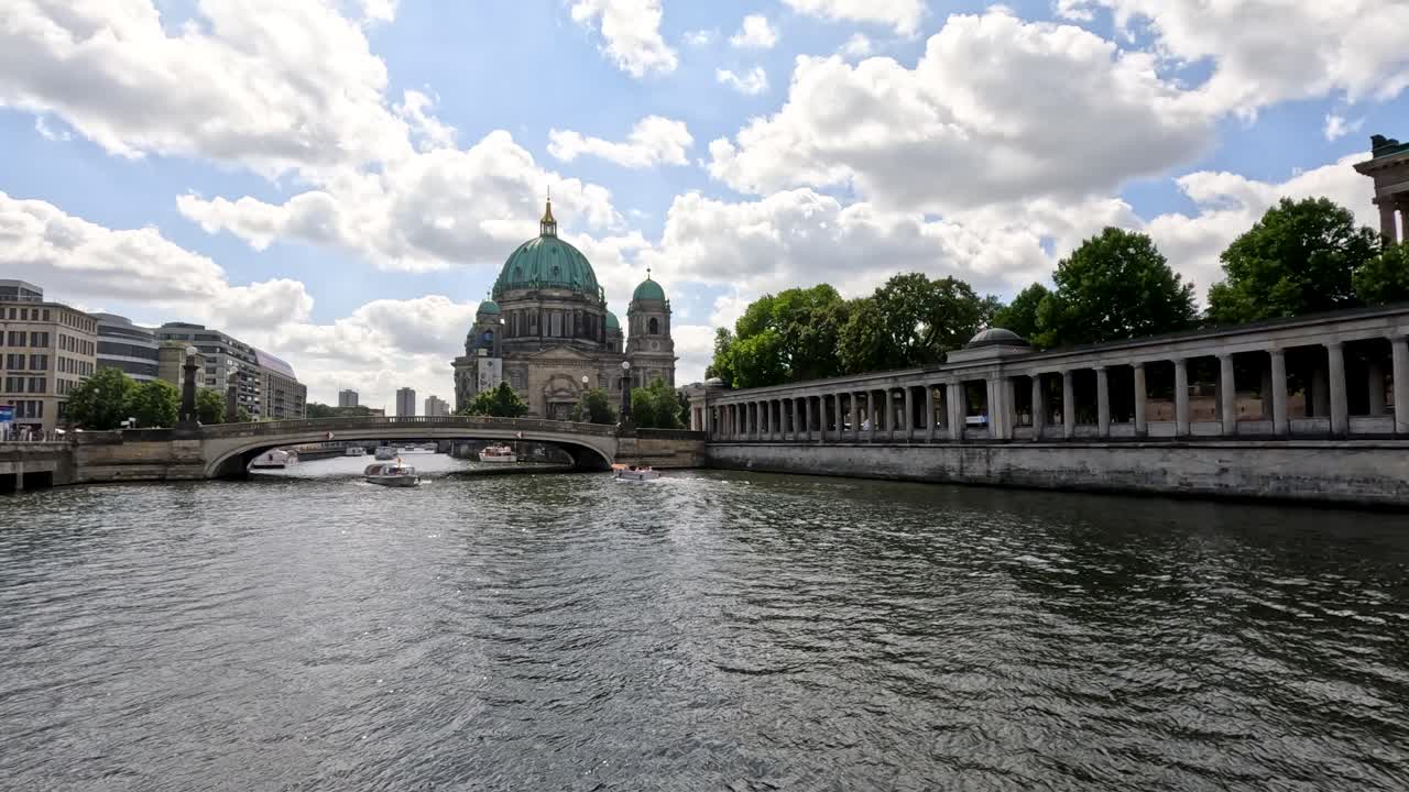 A boat travels smoothly on the Spree River in Berlin, approaching the historic cathedral under partly cloudy daylight. Wide, steady camera captures urban riverscape