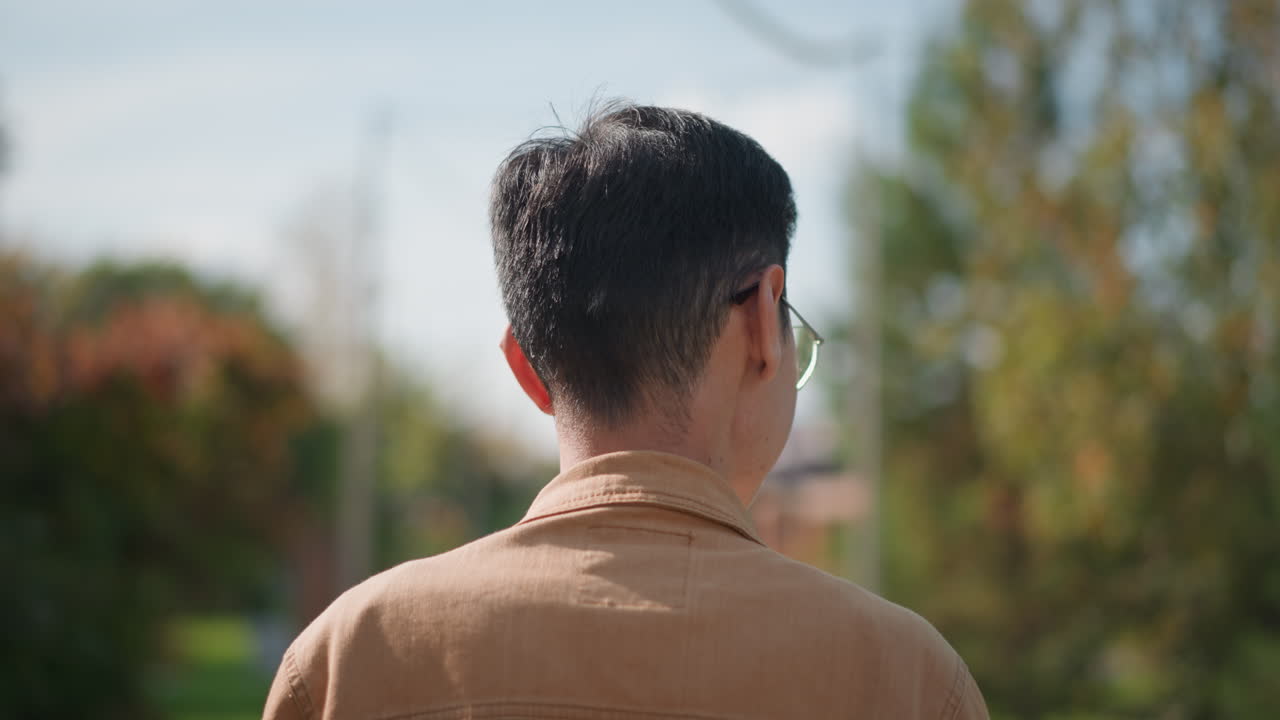 Giovane uomo su una strada alberata, che indossa occhiali e un maglione marrone, di spalle rispetto alla camera mentre guarda un murale distante e lo sfondo di un sobborgo verdeggiante, bokeh leggero che mette in risalto i colori autunnali, stile cinematografico