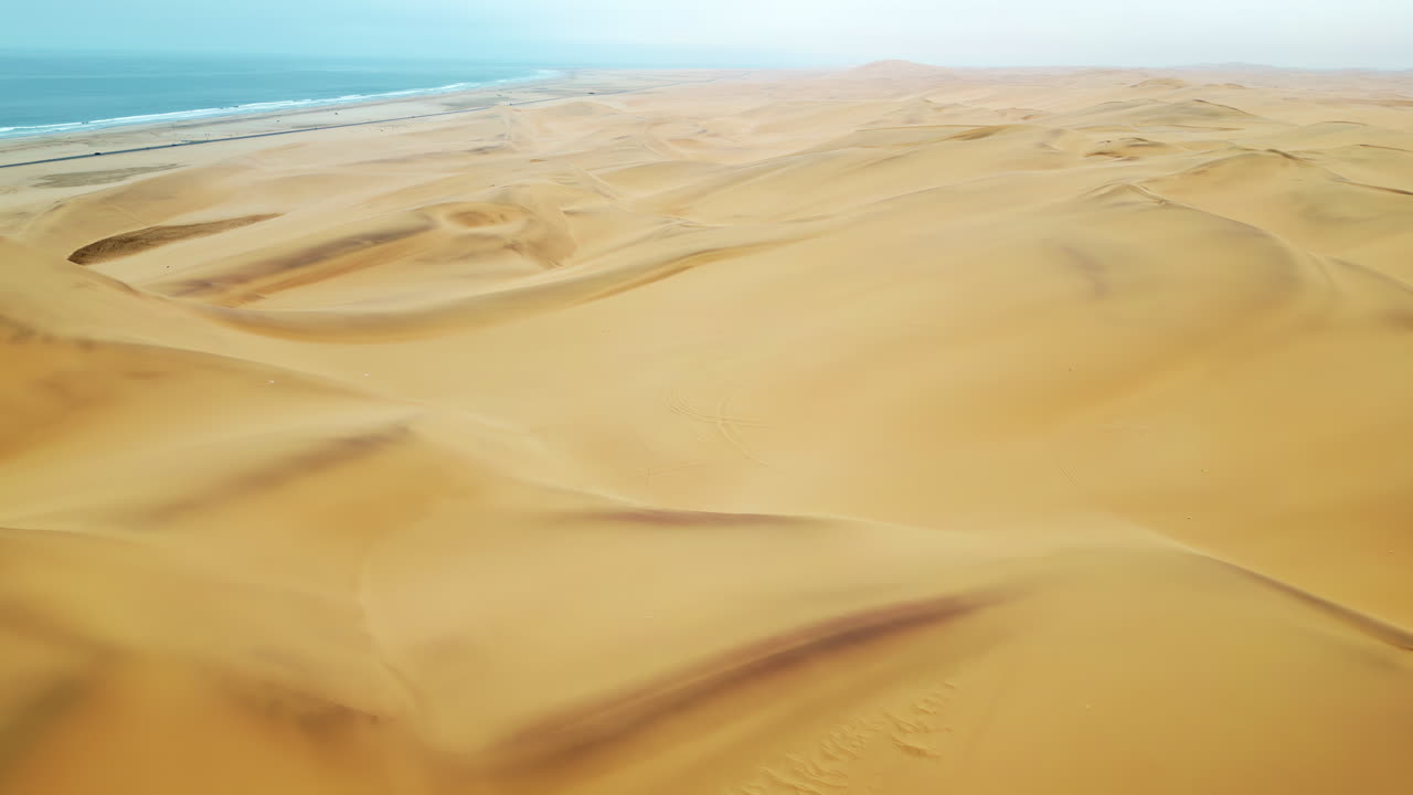 Vista aerea con drone delle dune di sabbia dorata del Deserto del Namib con tracce di pneumatici che attraversano le dune e l'Oceano Atlantico visibile in lontananza
