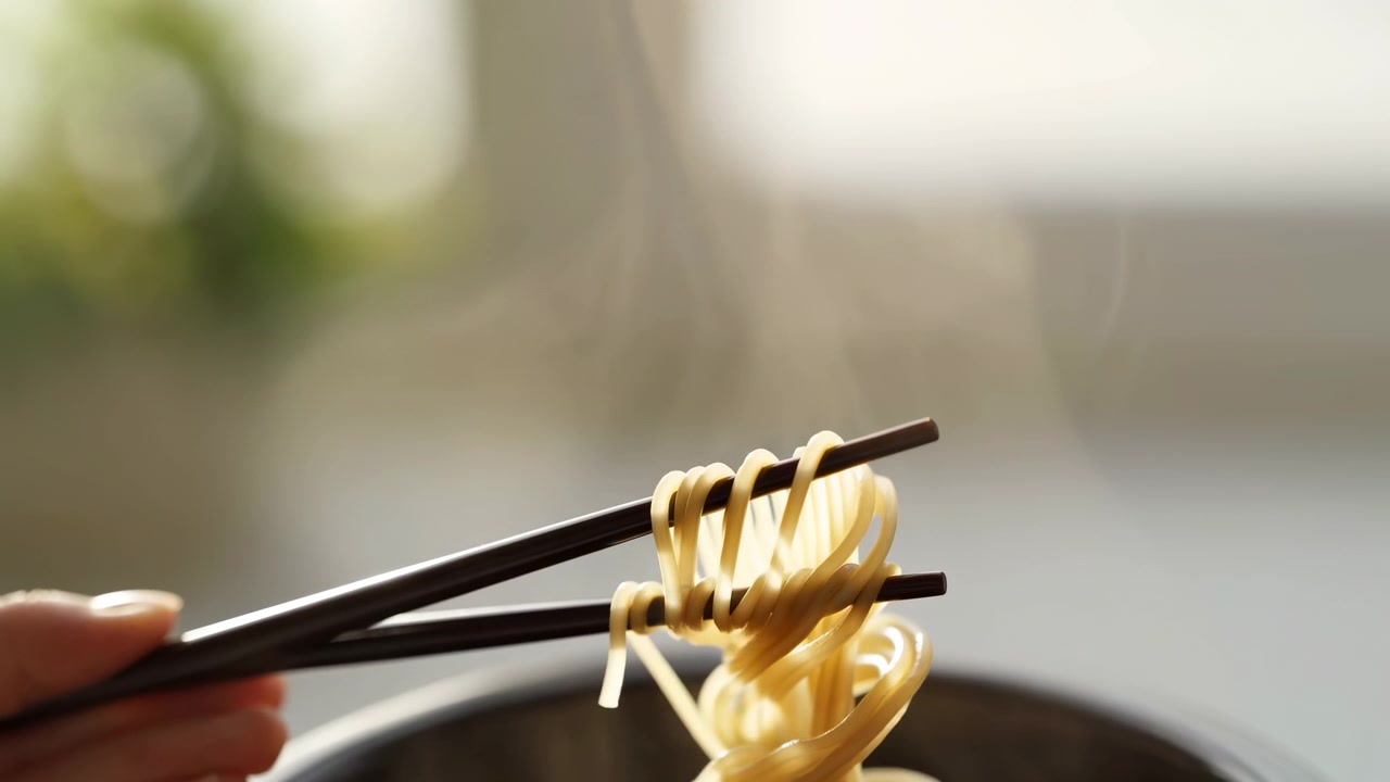 Spaghetti cotti avvolti su bacchette scure sopra il bordo della pentola, con presa della mano visibile sul bordo sinistro, vapore che sale, controluce caldo, sfocatura morbida e fili lucidi per un'invitante azione in cucina.