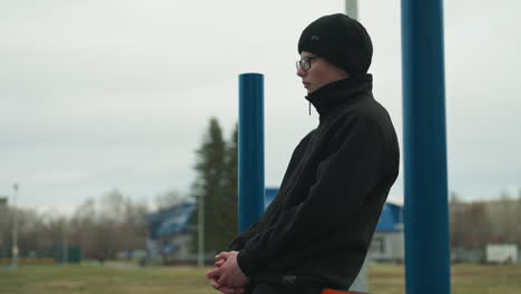 side view of a young boy dressed in a black outfit and glasses, sitting thoughtfully on a red iron bar, with a blur view of people in the in the field