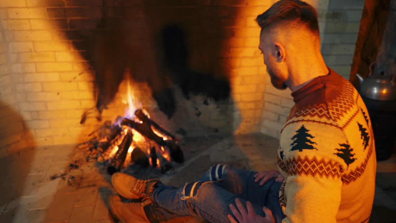 Young man warming his hands near the fireplace. Man in Christmas sweater and jeans sitting by the fire inside the house.