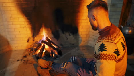 young man warming his hands near the fireplace. man in christmas sweater and jeans sitting by the fire inside the house.