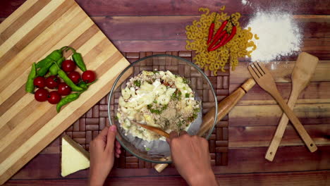 A-Top-view-of-marinating-the-macaroni-with-ingredients,-Marinated-macaroni-in-the-bowl,-raw-macaroni,-wooden-spoon,-wooden-spatula,-flour,-rolling-pin,-a-frozen-cheese-piece-and-chilies-on-the-table