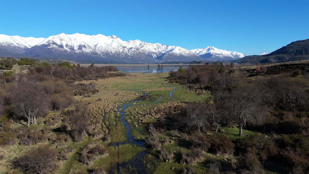 Flowing clear water meandering through a picturesque valley, surrounded by majestic snowy mountains under a bright sunny sky in Trevelin, Chubut, Argentina, aerial footage