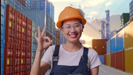 primer plano de una trabajadora asiática que usa gafas de protección y casco de seguridad sonriendo y mostrando un gesto de acuerdo a la cámara mientras está de pie en el almacén del patio de contenedores