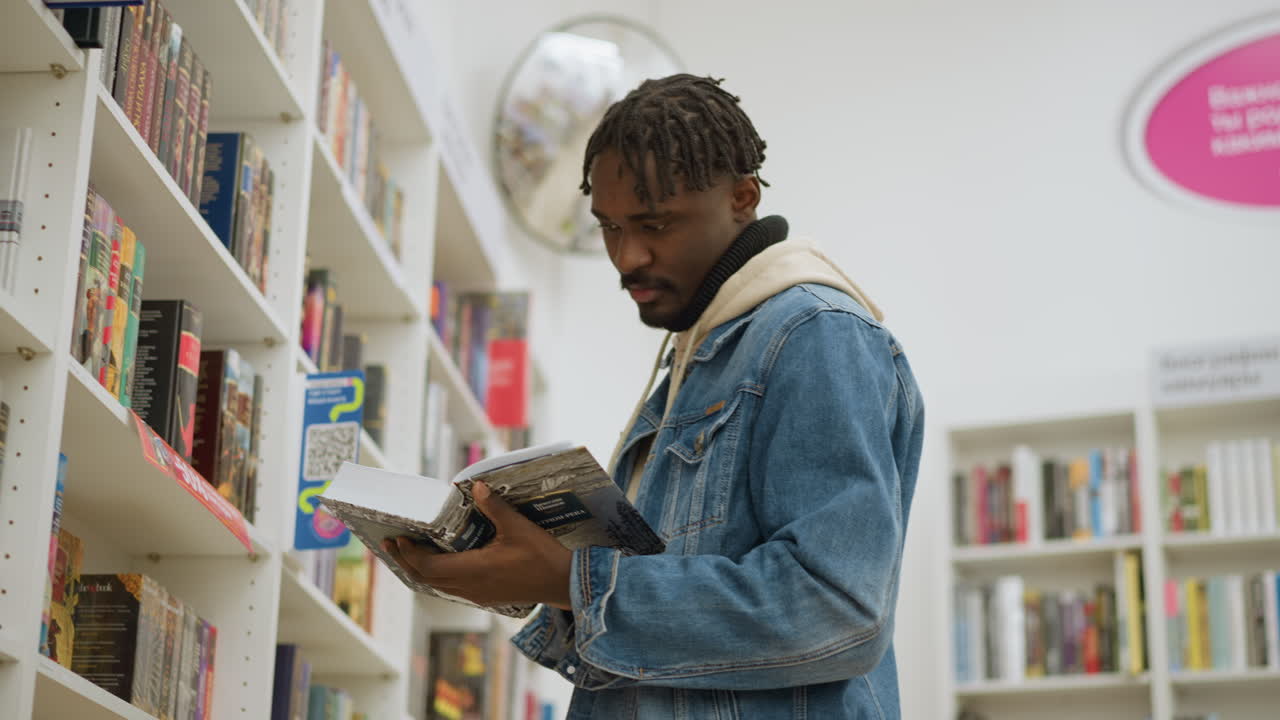 Student standing in modern library, holding open book in hands, examining pages with focus, surrounded by colorful book spines on shelves, exploring educational material in calm environment