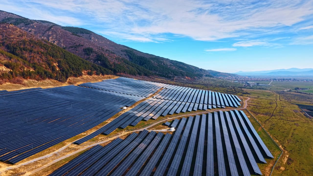 緑の丘陵と村の間に広がる太陽光発電所のドローンからの眺め。傾斜地に設置された角度のあるソーラーパネルの列、遠方には集落と広大な畑が広がる。