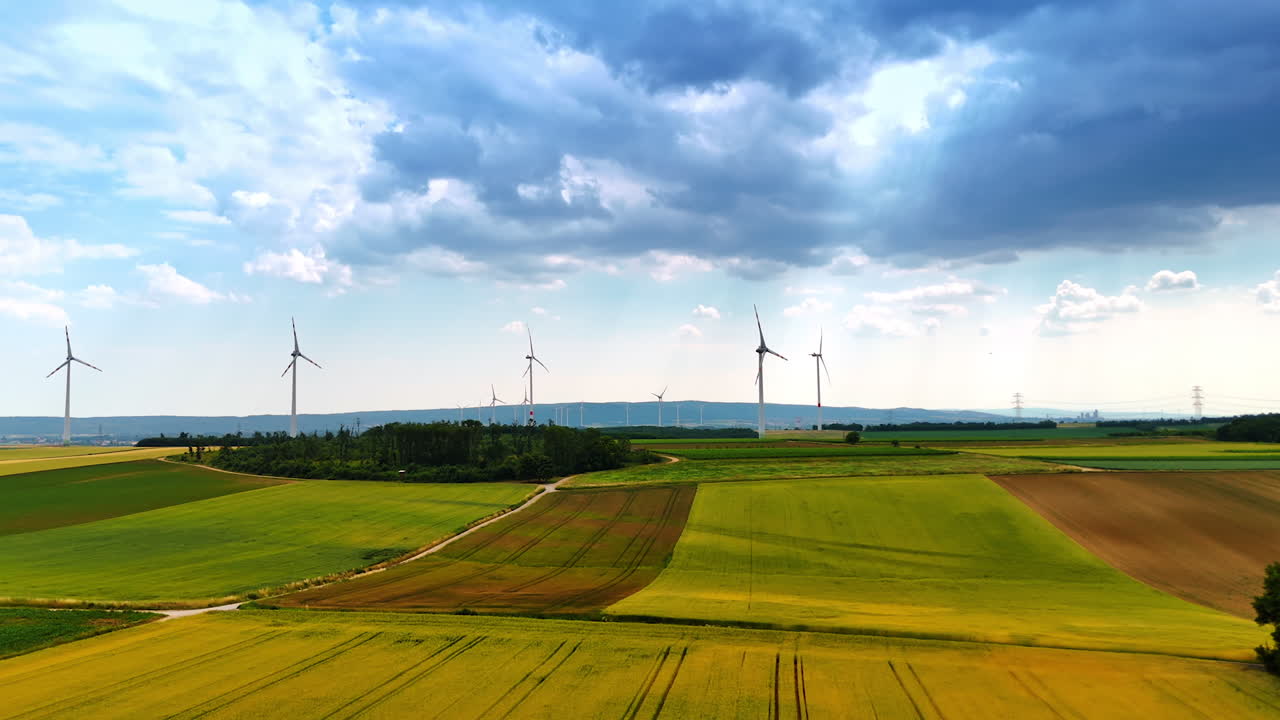Flight over the grassy meadows in the countryside on summer day. Multiple wind turbines rotate at backdrop. Green energy concept