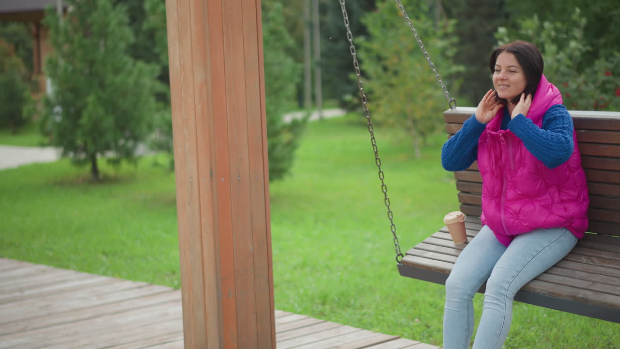 Donna su una panchina a dondolo in legno, che si sistema le cuffie e sorseggia un caffè, indossa un gilet rosa e un maglione blu con jeans chiari, si appoggia allo schienale e chiude gli occhi, leggera brezza tra gli alberi verdi, sentiero