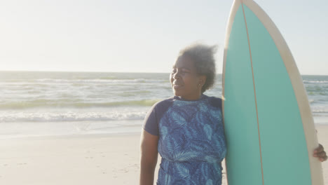 happy senior african american woman holding surfboard at beach, in slow motion, with copy space