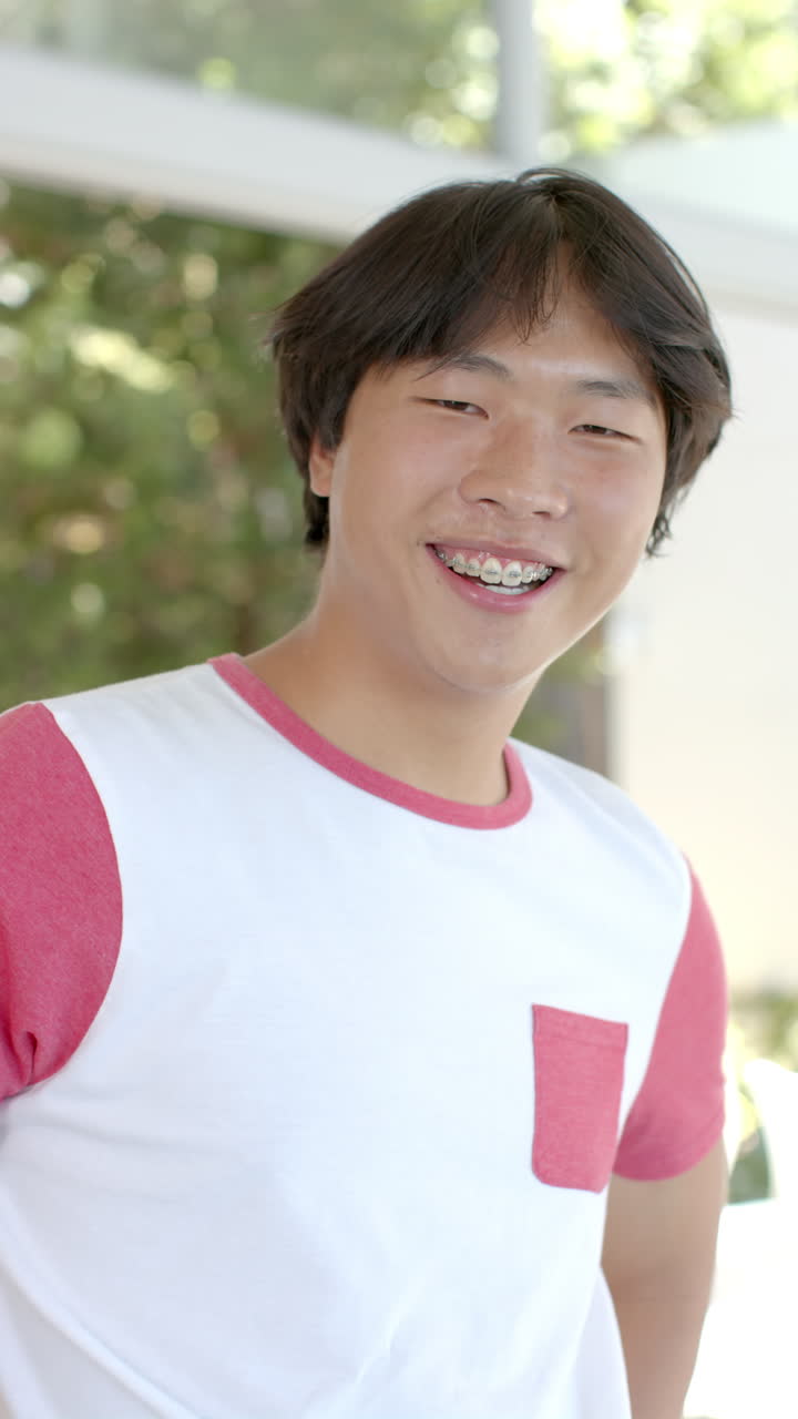 Vertical video: Smiling Asian teenage boy with braces wearing casual shirt at home, feeling happy
