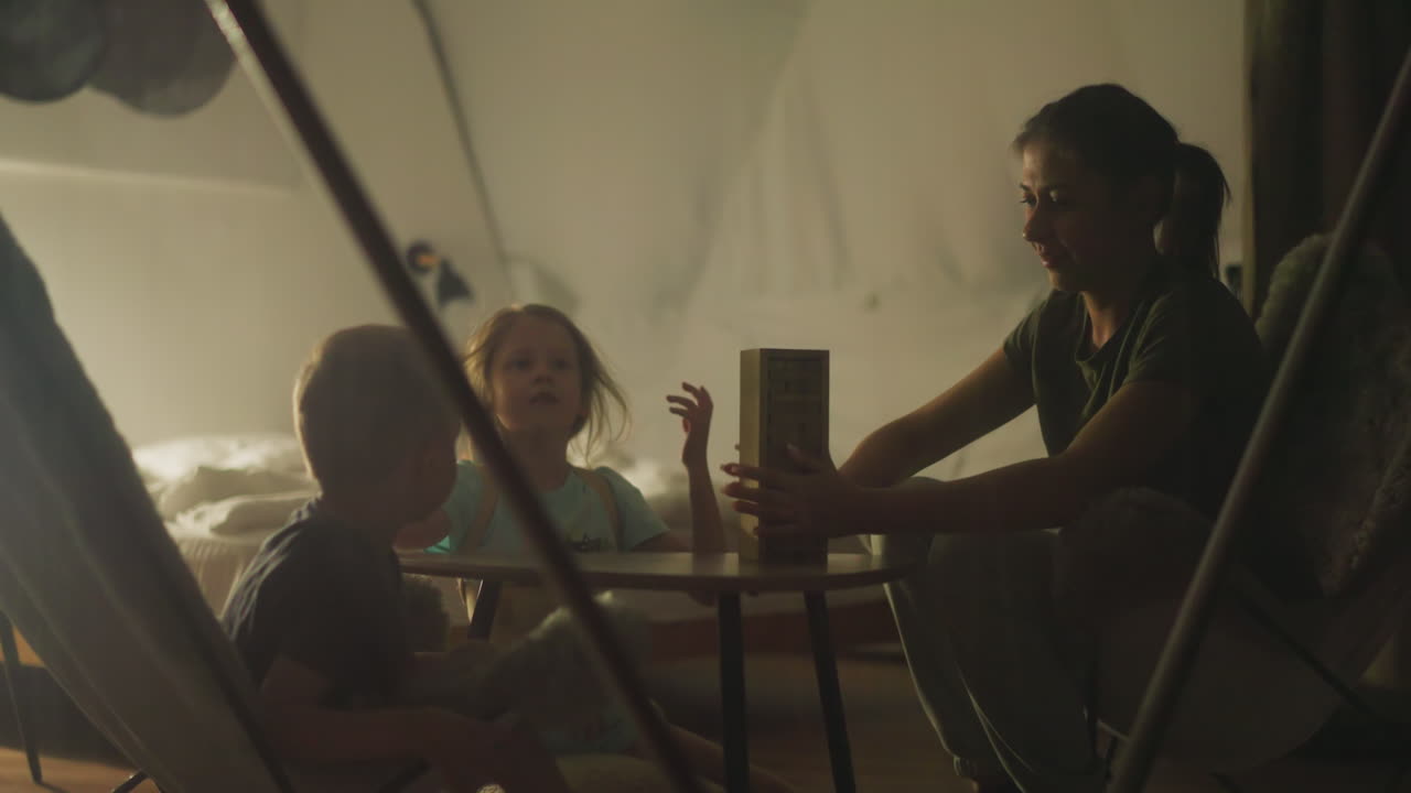 젊은 어머니는 글램핑 사이트 하우스 안의 관광 의자에 앉아있는 동안 보드 게임의 규칙을 아들과 딸에게 말합니다.