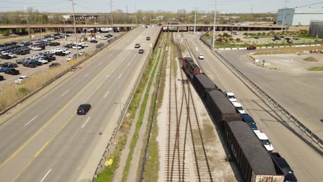 Train-hauling-coal-through-small-town-with-street-traffic,-aerial-follow-view