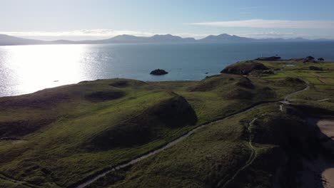 Aerial-view-Ynys-Llanddwyn-Welsh-hiking-island-with-shimmering-ocean-and-misty-Snowdonia-mountain-range-across-the-sunrise-skyline