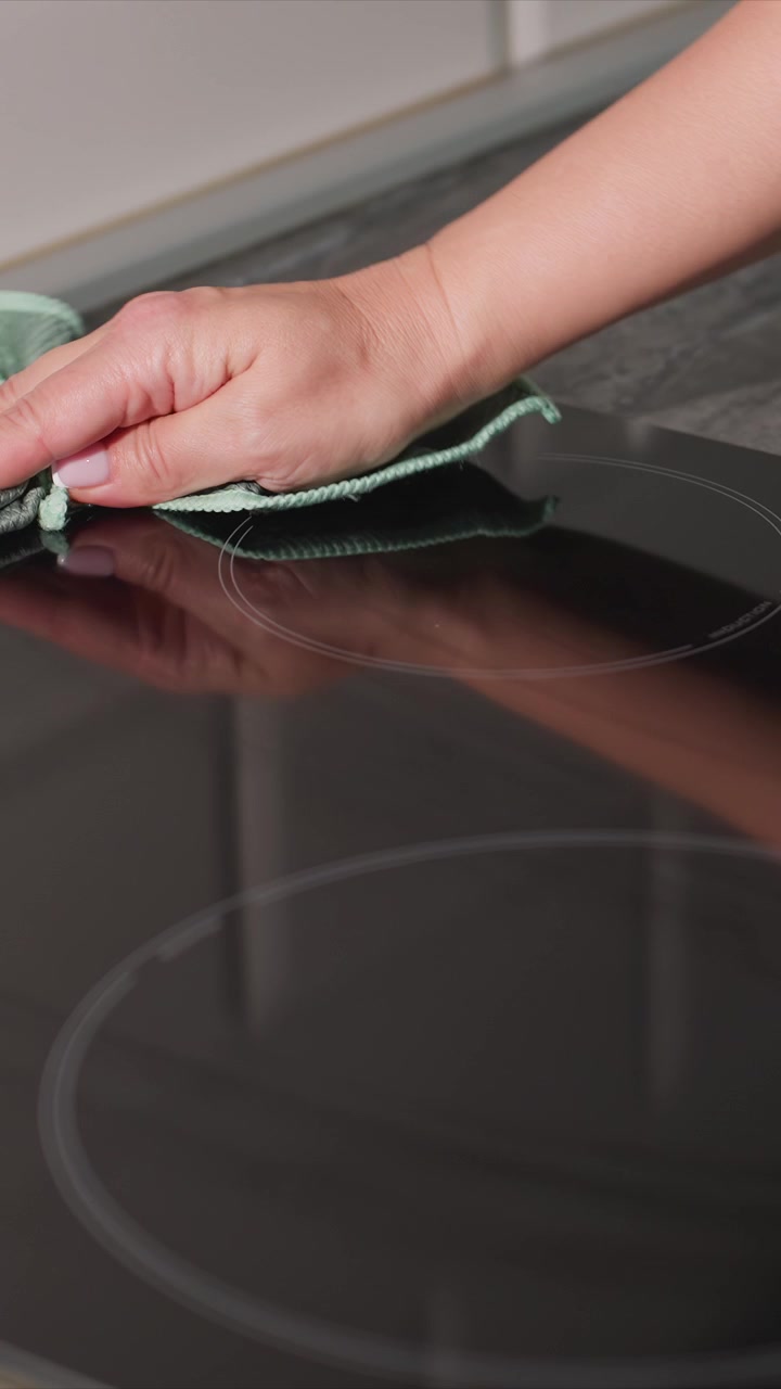 Primo piano di una mano con unghie ben curate che pulisce la superficie di un piano cottura a induzione usando un panno verde. Il vetro nero liscio riflette il movimento di pulizia, enfatizzando un movimento attento e costante.