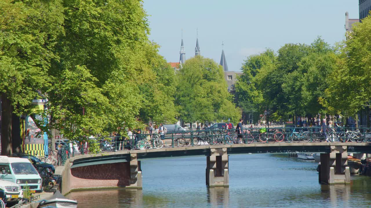 Bicyclists ride across a tree-lined bridge above a canal in central Amsterdam, daylight, wide shot