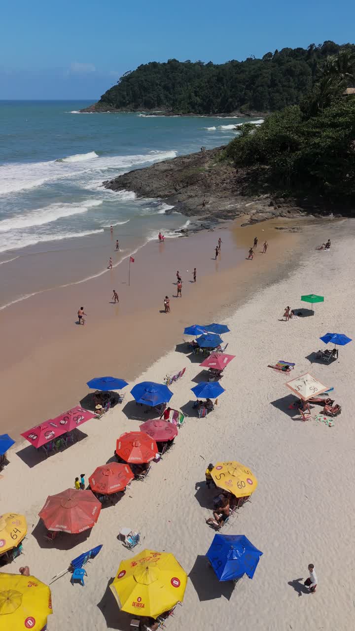 Drone descending Praia da Tiririca Beach Itacare Bahia Brazil, colorful umbrellas, people playing altinha, blue ocean, white waves, lush jungle, rocky coastline