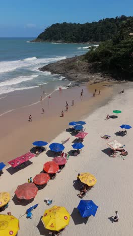 drone descending praia da tiririca beach itacare bahia brazil, colorful umbrellas, people playing altinha, blue ocean, white waves, lush jungle, rocky coastline